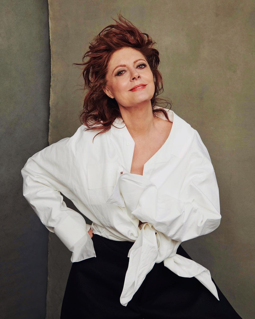 Portrait of Susan Sarandon wearing an oversized white button-down shirt and black pants against a textured green backdrop