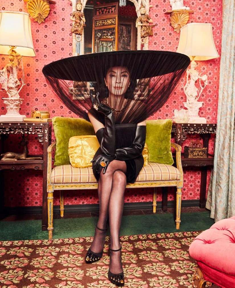 Michelle Yeoh seated on an ornate settee wearing an oversized black hat with veil, black leather gloves, and strappy heels.
