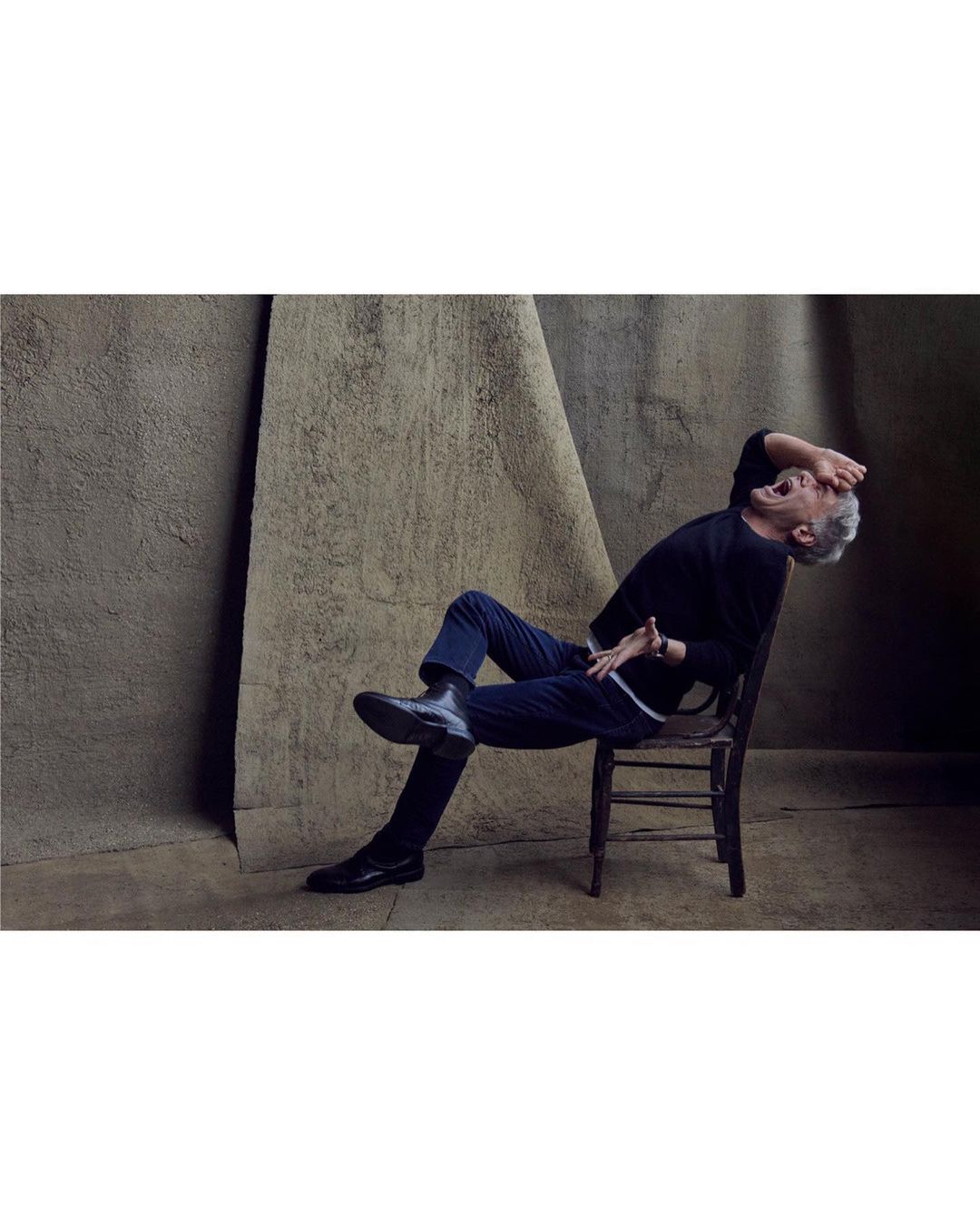 An adult male with grey hair sits on a wooden chair in a dark sweater and trousers against a textured neutral backdrop.