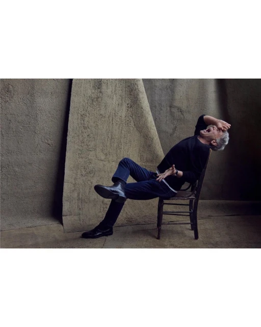 An adult male with grey hair sits on a wooden chair in a dark sweater and trousers against a textured neutral backdrop.