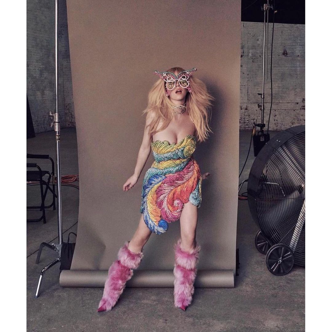 Fashionable woman in a strapless feather dress and decorative eyewear strikes a pose in a photo studio with vintage fan and lighting.