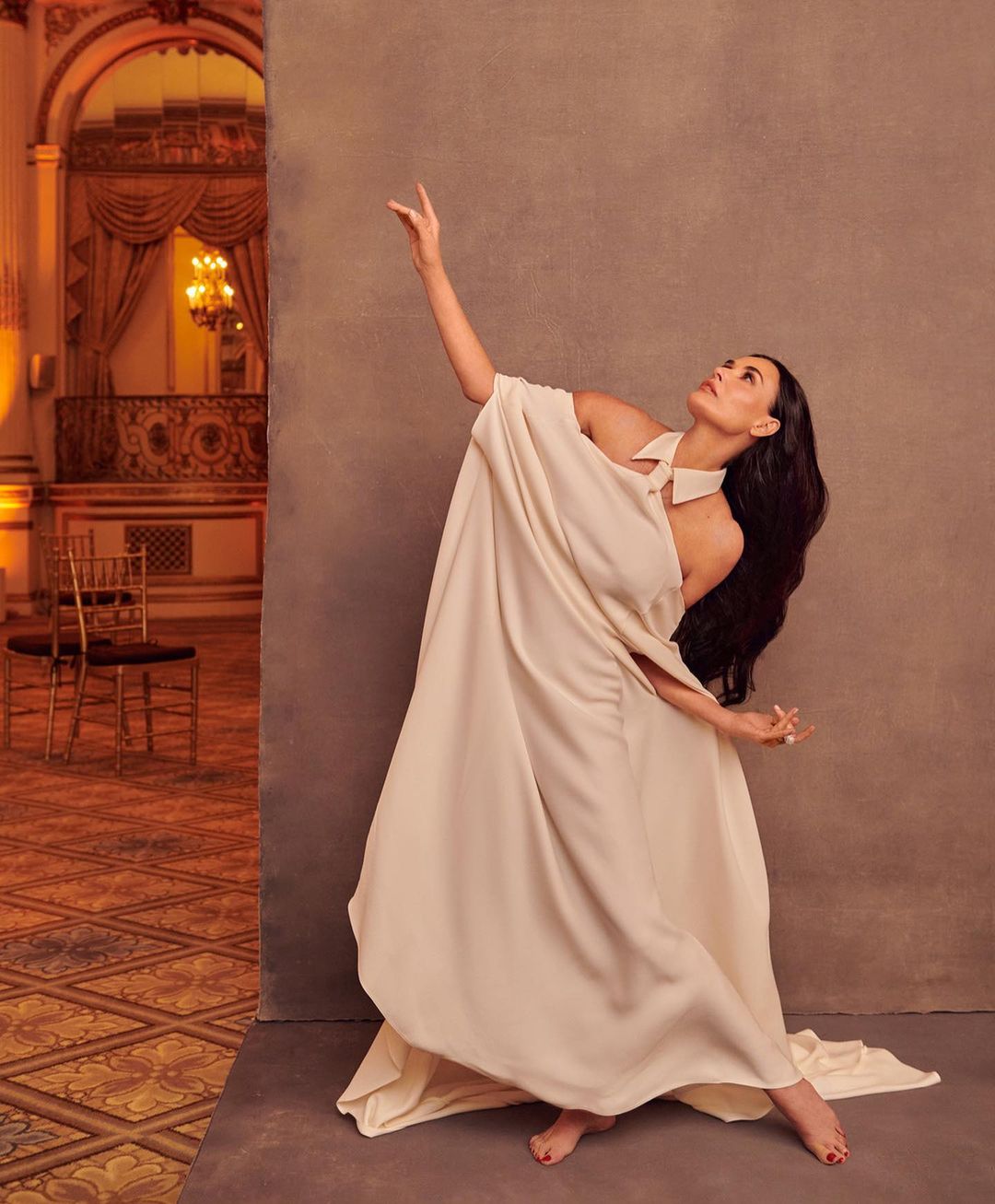 Woman in flowing white dress strikes a pose in a ballroom. Warm lighting accentuates the elegance of her attire and the ornate setting.