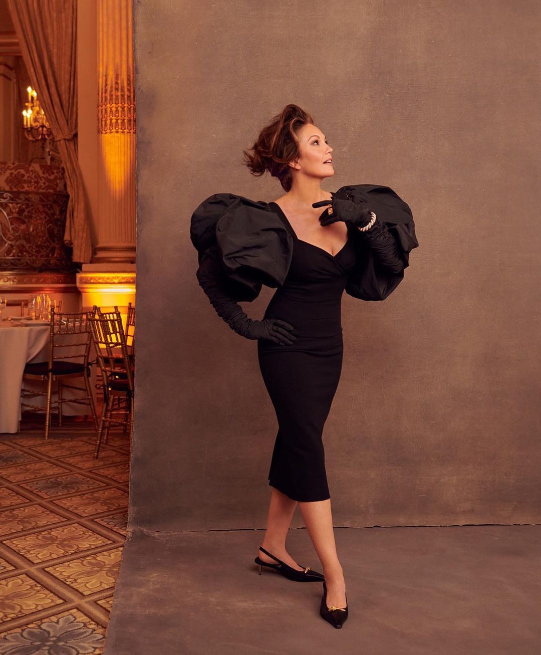 Elegant woman poses in a black dress with dramatic sleeves and gloves, set against a textured backdrop with a classic ballroom visible.
