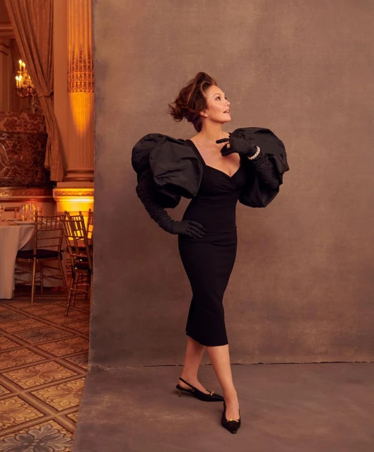 Elegant woman poses in a black dress with dramatic sleeves and gloves, set against a textured backdrop with a classic ballroom visible.