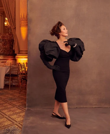 Elegant woman poses in a black dress with dramatic sleeves and gloves, set against a textured backdrop with a classic ballroom visible.