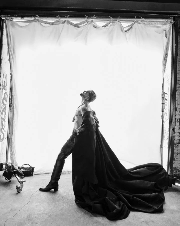 Monochrome portrait of a man in a sweeping cape and tall boots posing in an open industrial doorway by photographer Ruven Afanador.