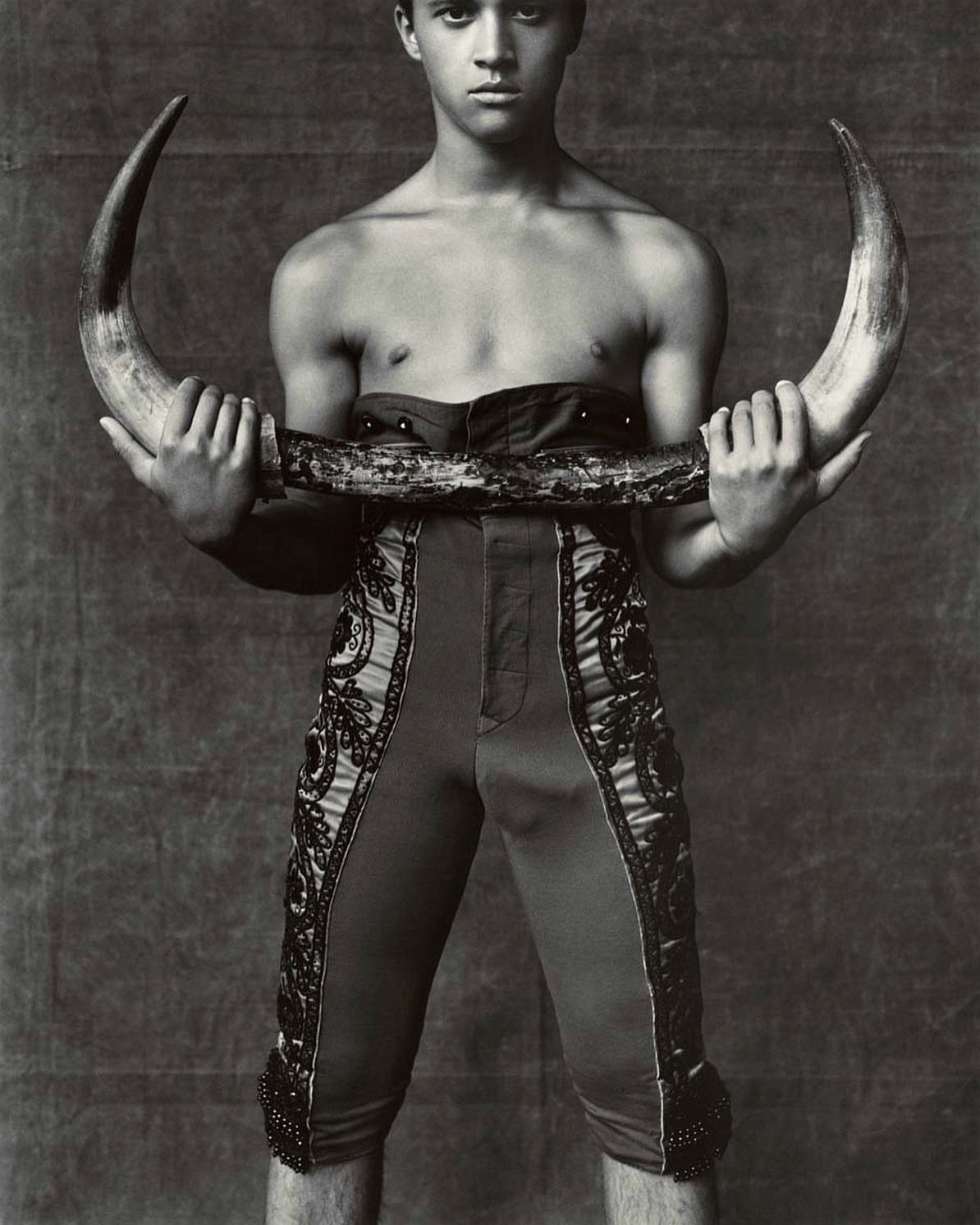 Monochromatic portrait of a young man holding bull horns, wearing traditional torero-style pants, against a textured background.