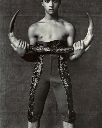 Monochromatic portrait of a young man holding bull horns, wearing traditional torero-style pants, against a textured background.