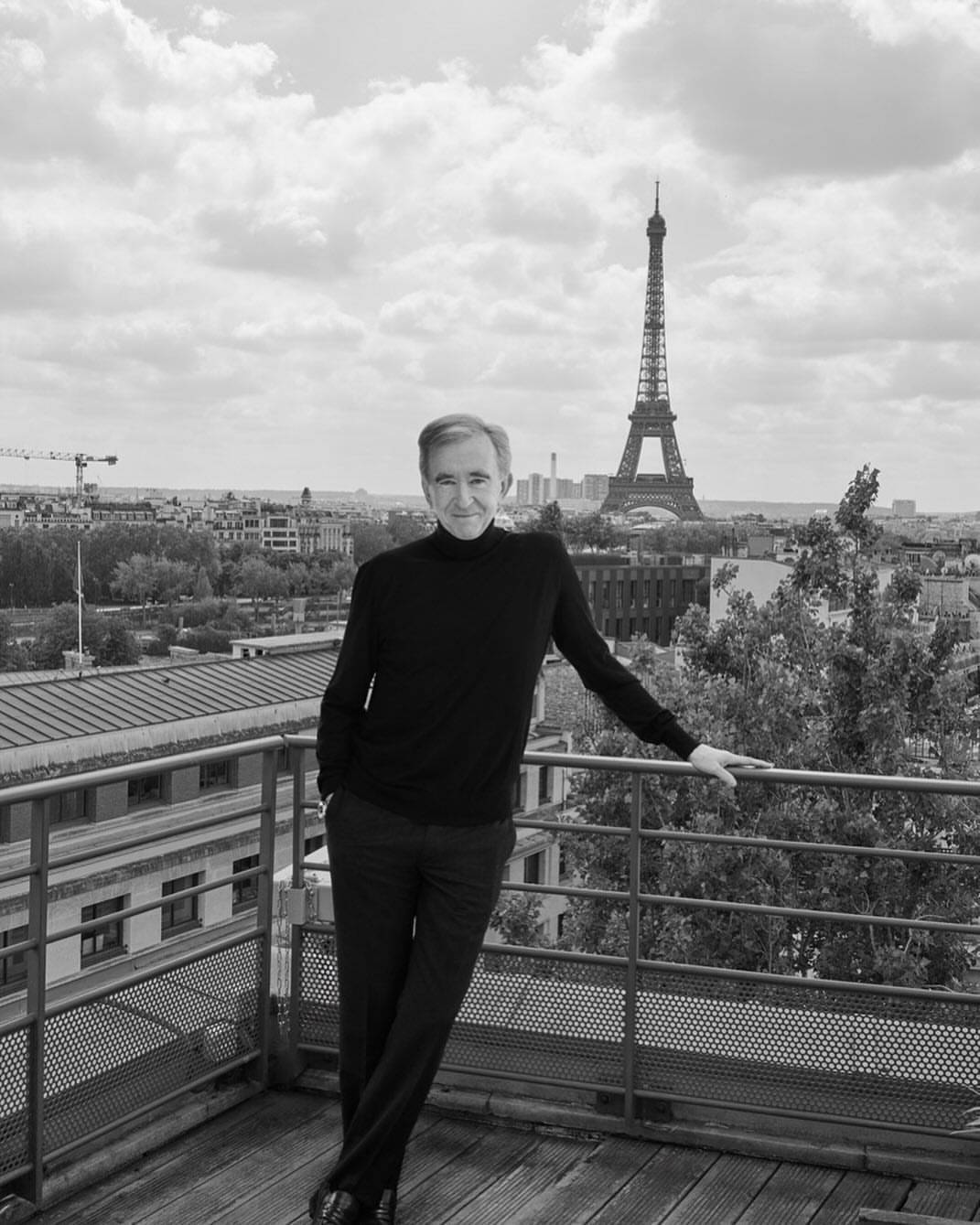 Black and white portrait of a man in a black turtleneck and pants leaning against a metal railing with the Eiffel Tower in background.