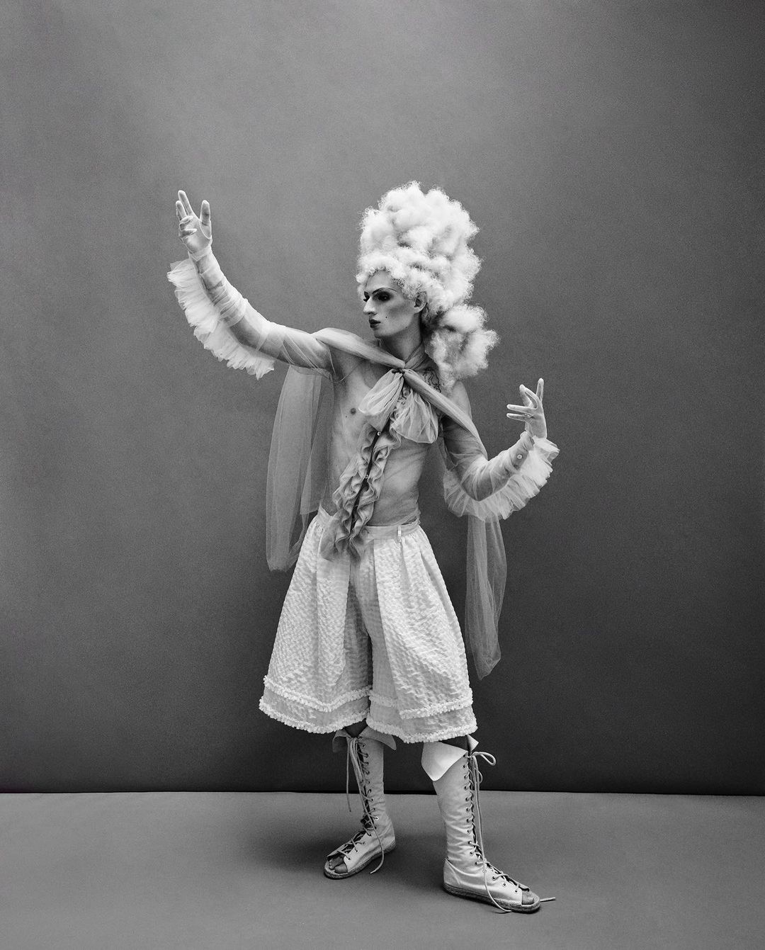 Black and white studio shot of a person in vintage clothing with a frilled cape, high-waisted shorts, knee-high laced boots, and large white wig.