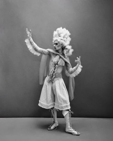 Black and white studio shot of a person in vintage clothing with a frilled cape, high-waisted shorts, knee-high laced boots, and large white wig.