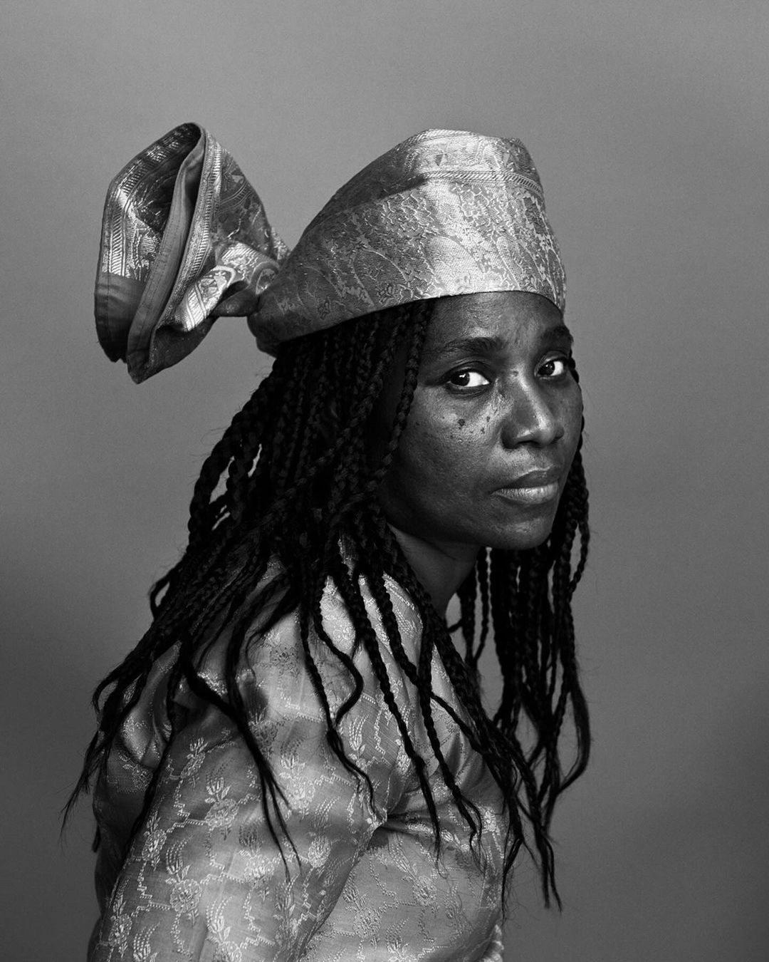 Portrait of an adult woman with dark skin, braided hair, and traditional headwear, posing against a grey studio backdrop in black and white.