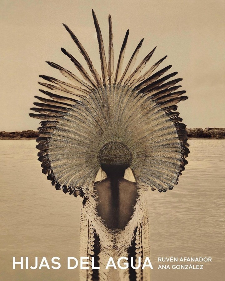 Woman wearing feathered headdress and fringe garment near river, soft light, earthen tones. Indigenous fashion editorial, environmental portrait.