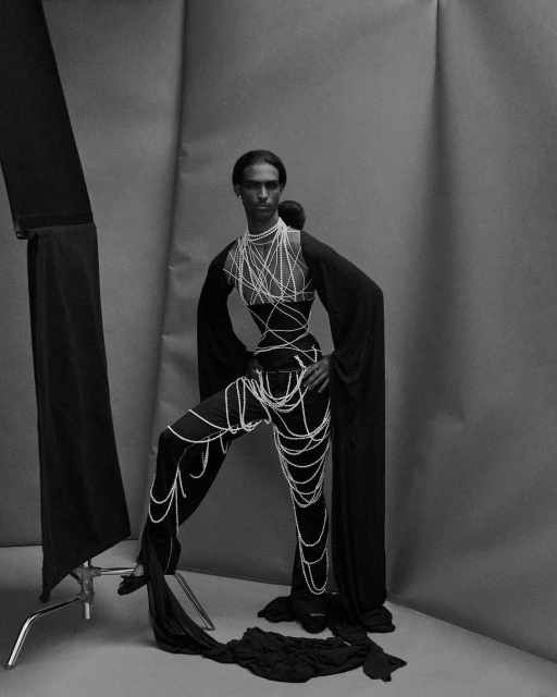 Monochrome fashion shot of a slender adult, draped in pearl-like strands, standing in a photo studio with soft lighting and dark clothing.
