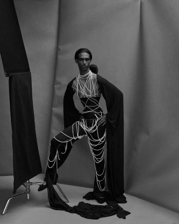 Monochrome fashion shot of a slender adult, draped in pearl-like strands, standing in a photo studio with soft lighting and dark clothing.