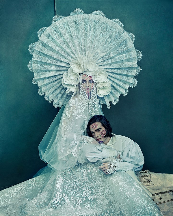 Woman in elaborate lace headpiece poses with man in ruffled period clothing with decorative facial makeup in a studio setting.