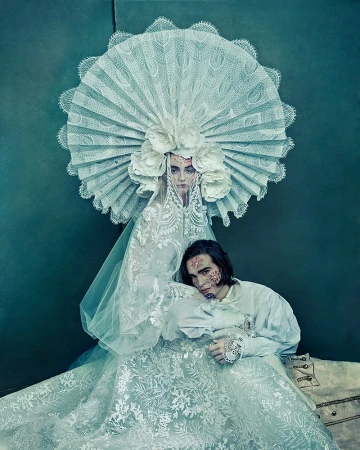 Woman in elaborate lace headpiece poses with man in ruffled period clothing with decorative facial makeup in a studio setting.