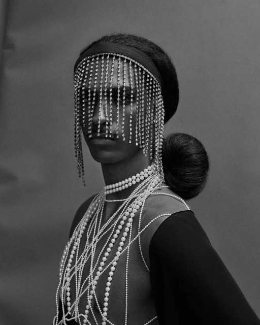Portrait of a woman with a beaded headpiece and multiple strands of pearls, set against a grey backdrop. Black and white photography.