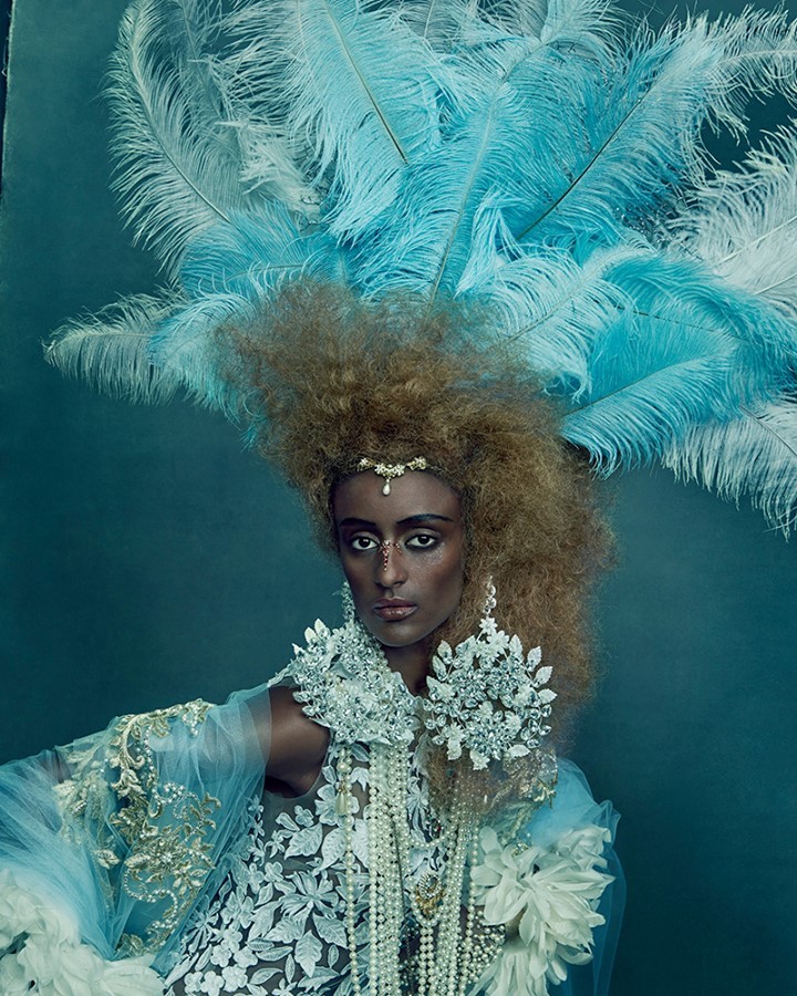 Portrait of a person with medium-dark skin in elaborate white and blue feathered headdress and jewelry against teal background.