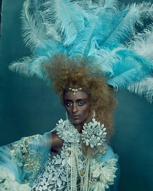 Portrait of a person with medium-dark skin in elaborate white and blue feathered headdress and jewelry against teal background.