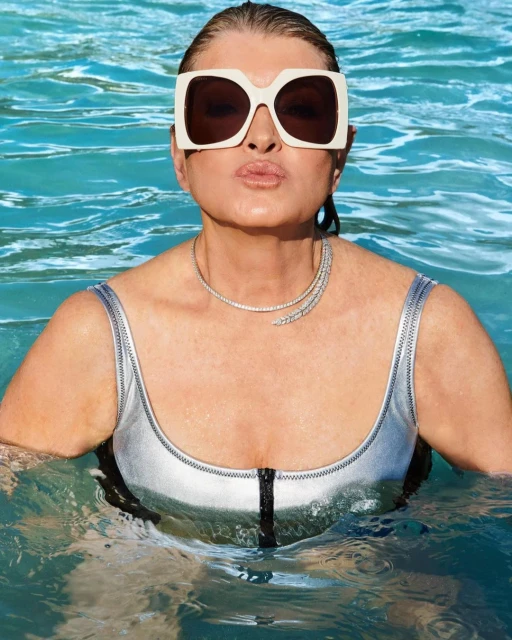 A woman with a silver swimsuit, sunglasses, and necklace is in the water looking at the camera with a kissy face expression.