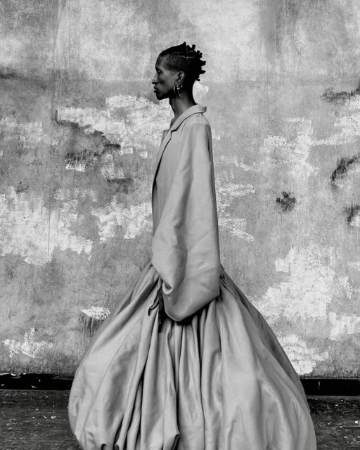 Monochrome portrait of a woman with braided hair in side profile, wearing an oversized dress, against a textured wall.