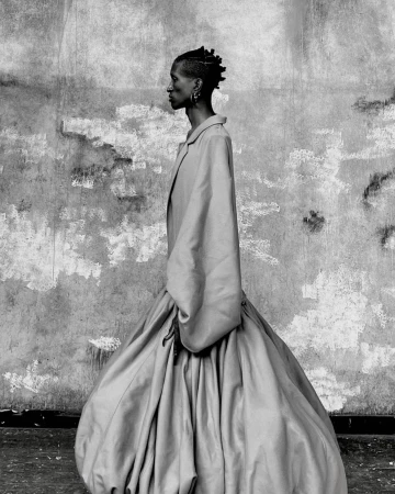 Monochrome portrait of a woman with braided hair in side profile, wearing an oversized dress, against a textured wall.