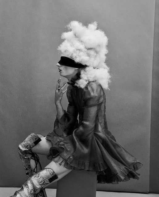 Fashion editorial shot of a person with an ornate blonde wig, wearing a red dress and snake-print boots, sitting in a photo studio.