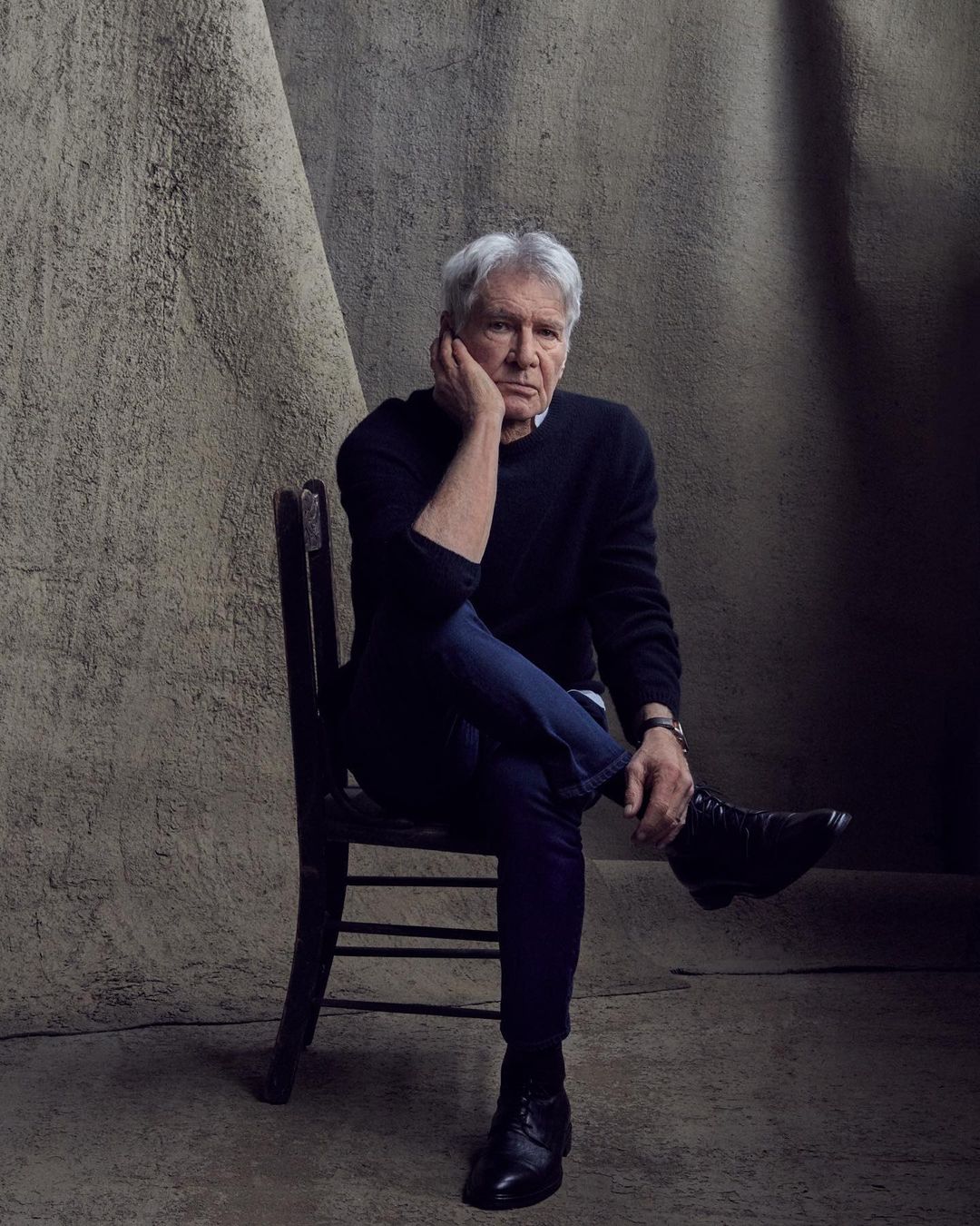 Portrait of Harrison Ford seated against a textured backdrop, wearing a dark sweater and blue jeans in a photo studio setting.