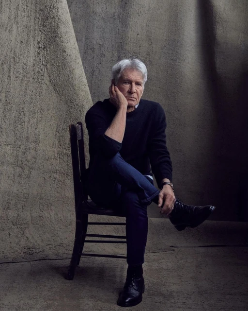 Portrait of Harrison Ford seated against a textured backdrop, wearing a dark sweater and blue jeans in a photo studio setting.