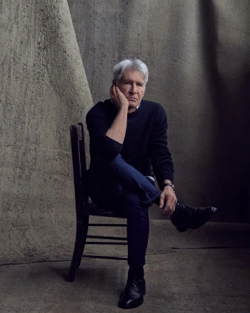 Portrait of Harrison Ford seated against a textured backdrop, wearing a dark sweater and blue jeans in a photo studio setting.