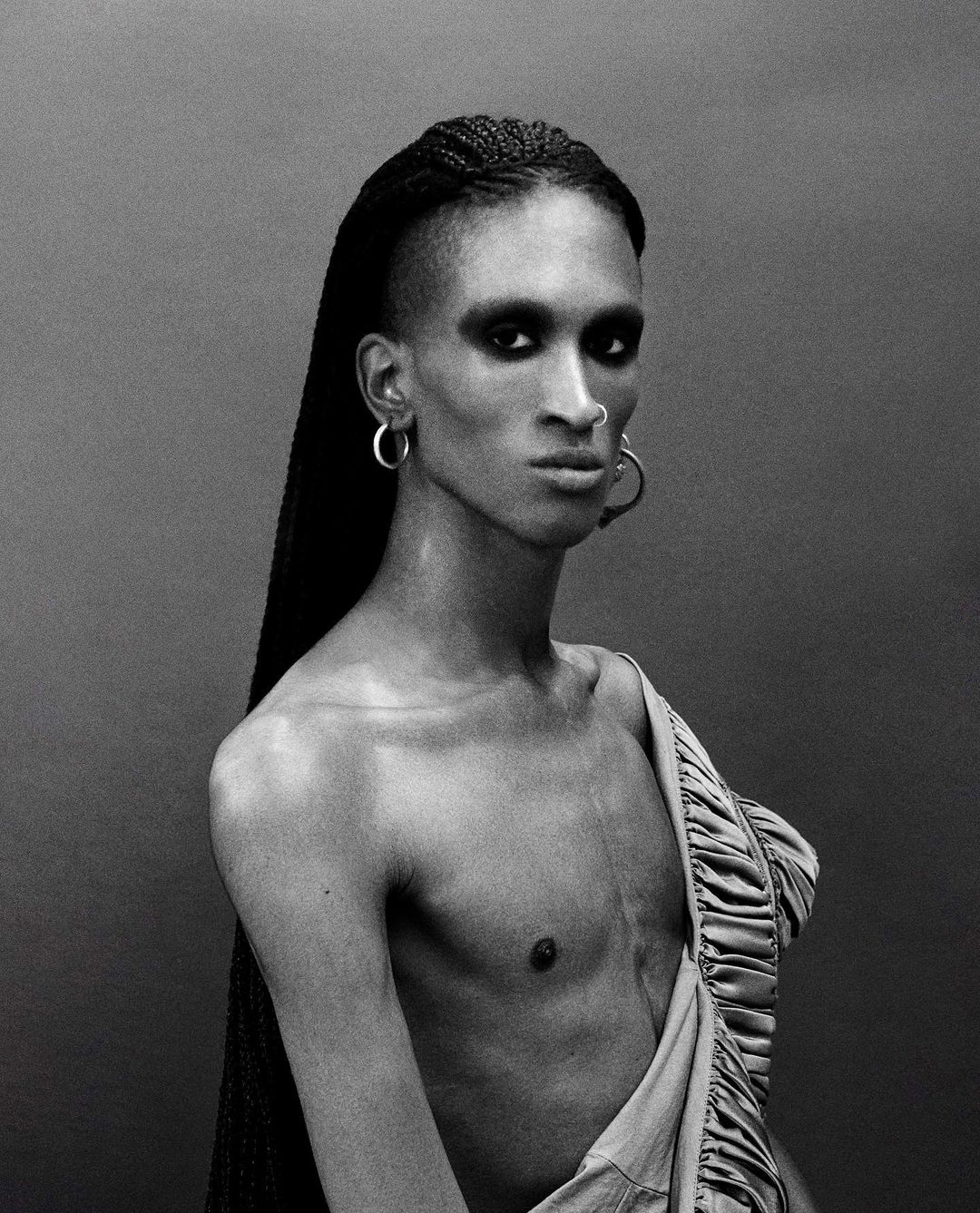 Striking monochrome portrait of a gender-neutral person with braided hair, dramatic eye makeup, and exposed torso, wearing hoop earrings.
