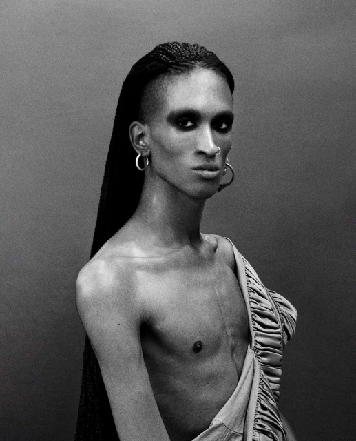 Striking monochrome portrait of a gender-neutral person with braided hair, dramatic eye makeup, and exposed torso, wearing hoop earrings.