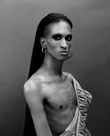 Striking monochrome portrait of a gender-neutral person with braided hair, dramatic eye makeup, and exposed torso, wearing hoop earrings.