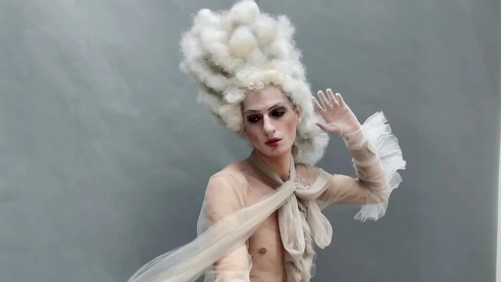 Individual in theatrical makeup and oversized white wig posing against a grey backdrop with a tulle dress and ruffled sleeves.
