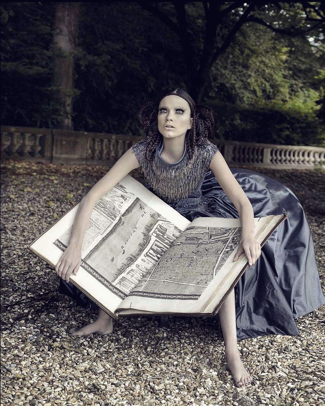 A young adult woman sits outdoors holding open a large book with architectural renderings, posing in a dark dress, in an artistic outdoor portrait.