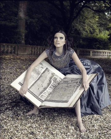A young adult woman sits outdoors holding open a large book with architectural renderings, posing in a dark dress, in an artistic outdoor portrait.