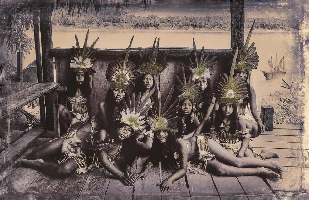 Group portrait of ten Indigenous people wearing feathered headdresses and tribal attire, posing indoors on a wooden structure with river view.