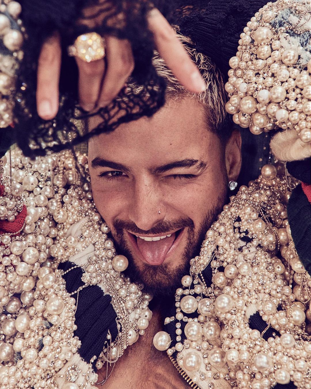 Close-up of a man with light brown hair and a goatee winking, wearing a pearl-covered jacket and holding up a diamond ring.