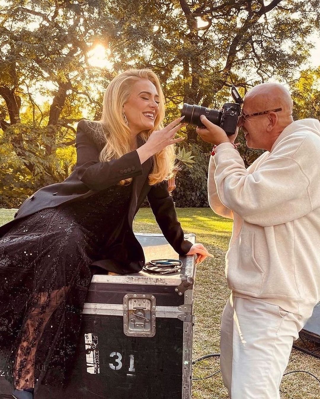 Adele poses with a photographer outdoors, wearing a black blazer and dress. She is smiling and making a hand gesture.
