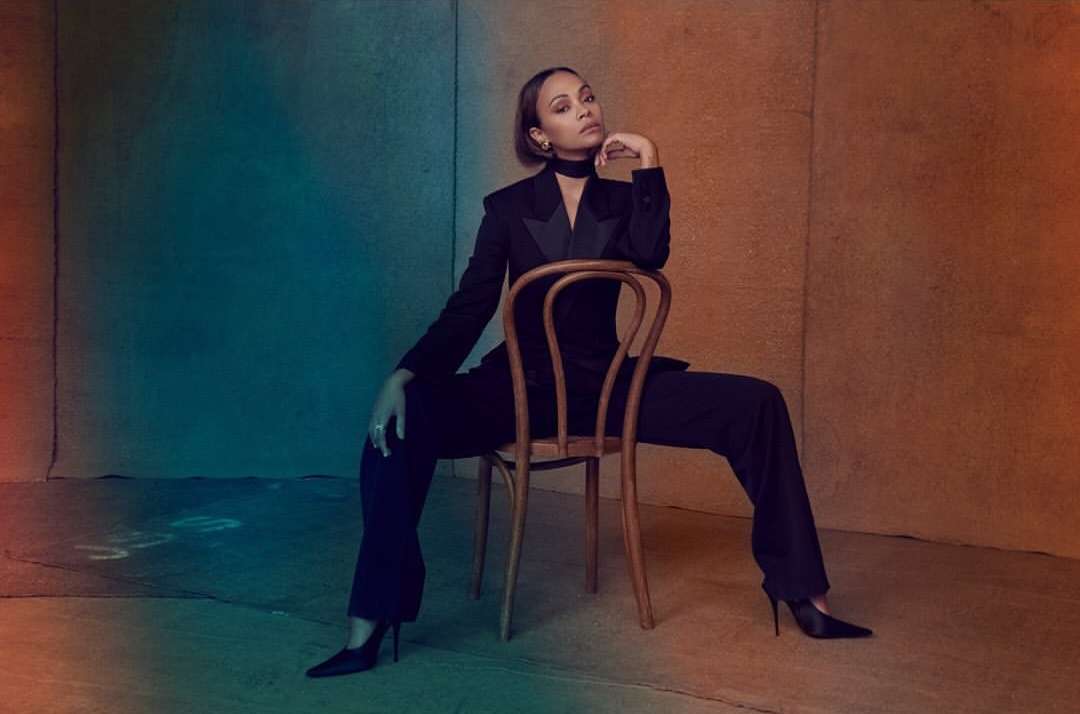 Zoe Saldana sits in a studio in an unbuttoned black suit with black high heels, posing with her hand near her face.