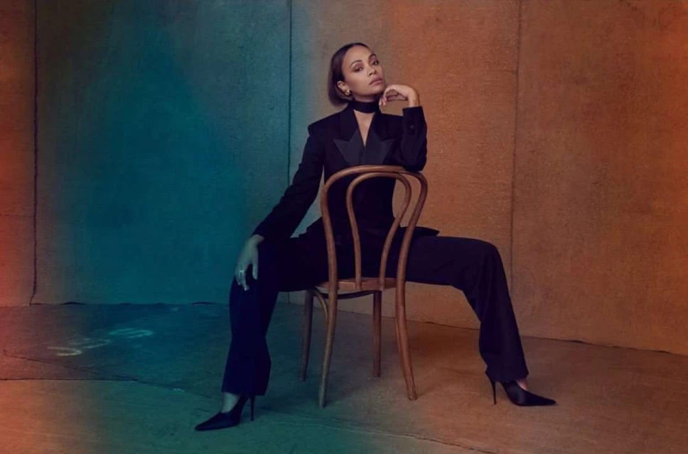 Zoe Saldana sits in a studio in an unbuttoned black suit with black high heels, posing with her hand near her face.