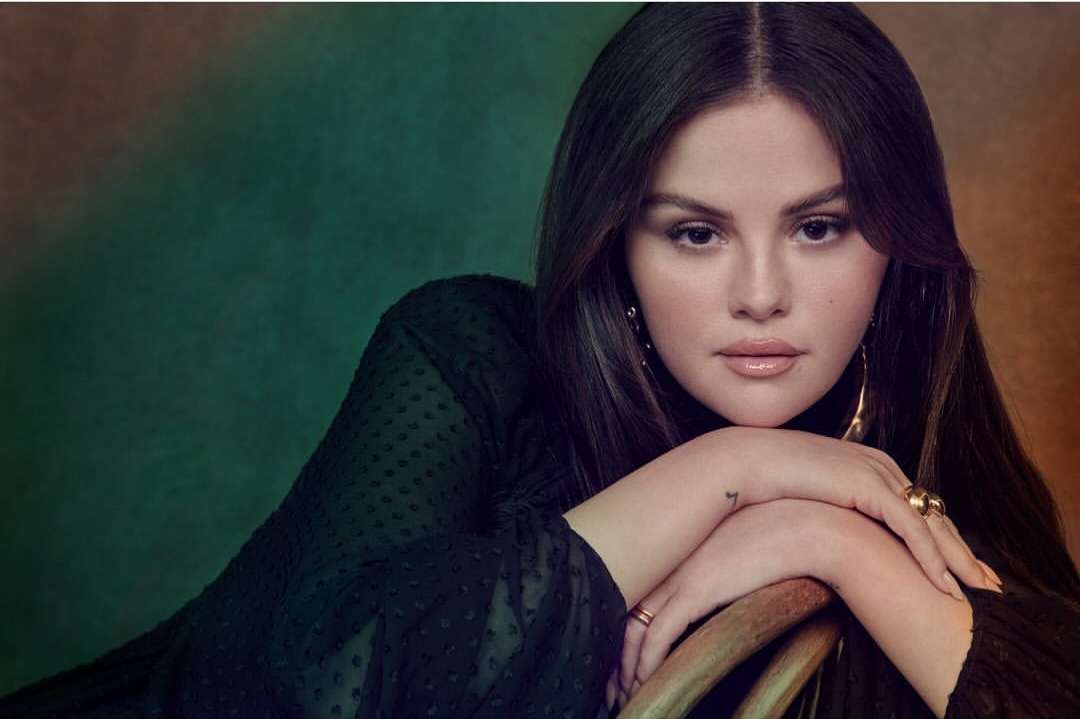 Close-up portrait of a woman with long dark hair in a black top, resting her arms on a chair, set against a blurred green and brown background.