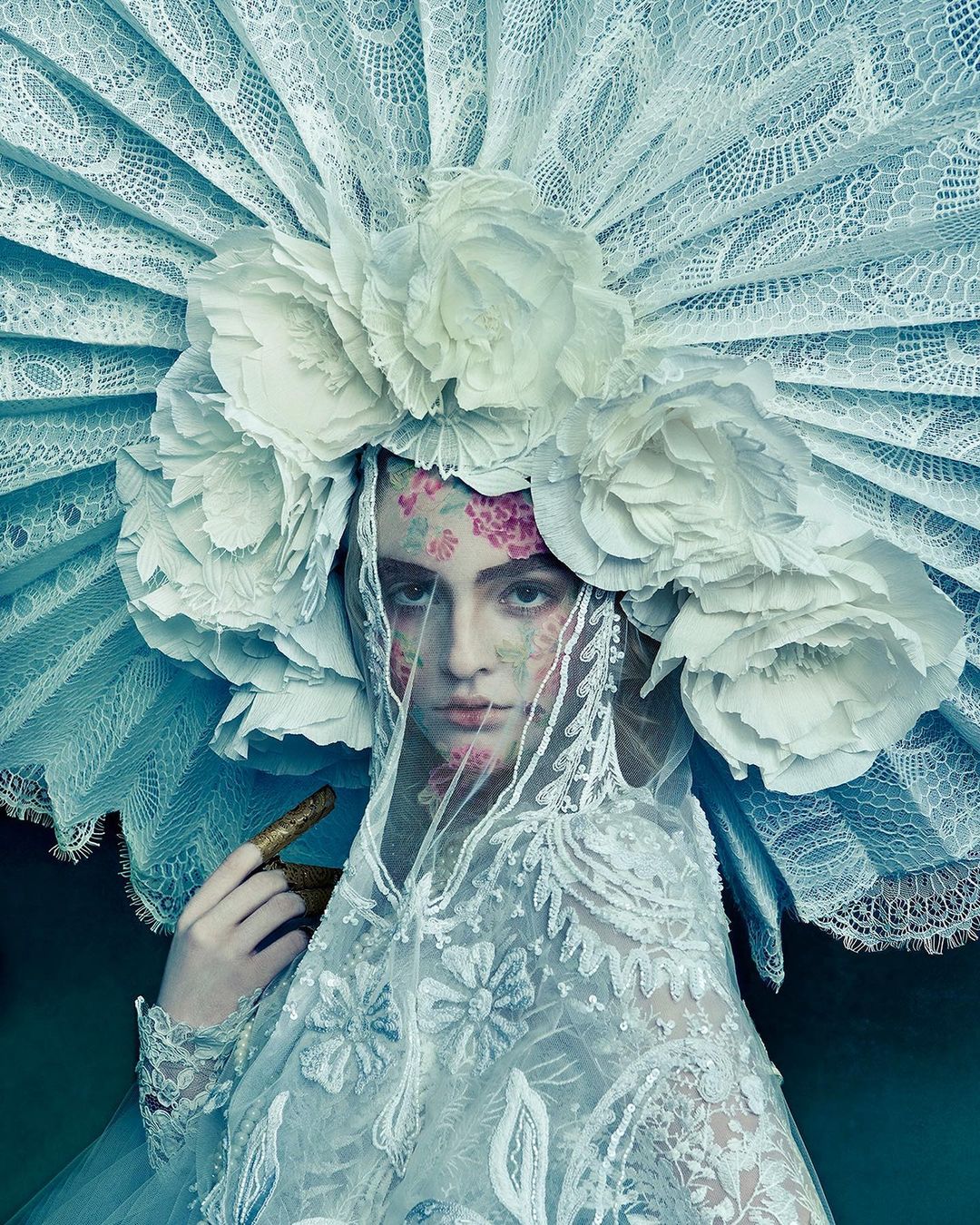 Woman with floral face art poses with a decorative white lace headpiece and veil in a portrait in an indoor setting.