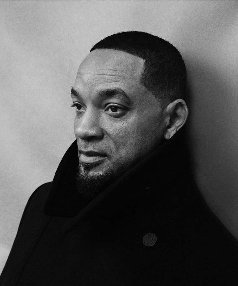 Black and white portrait of a man with a goatee wearing a dark coat against a neutral backdrop in a studio setting.