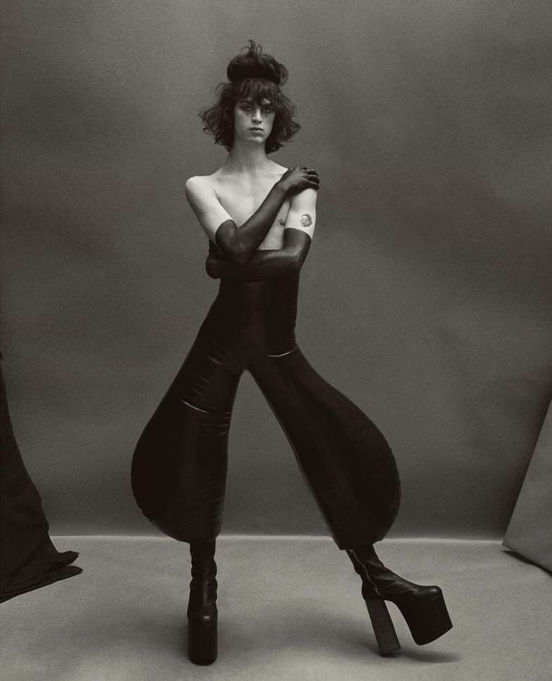 Black-and-white portrait of a person in a black bodysuit and platform boots posing in a studio against a neutral background.