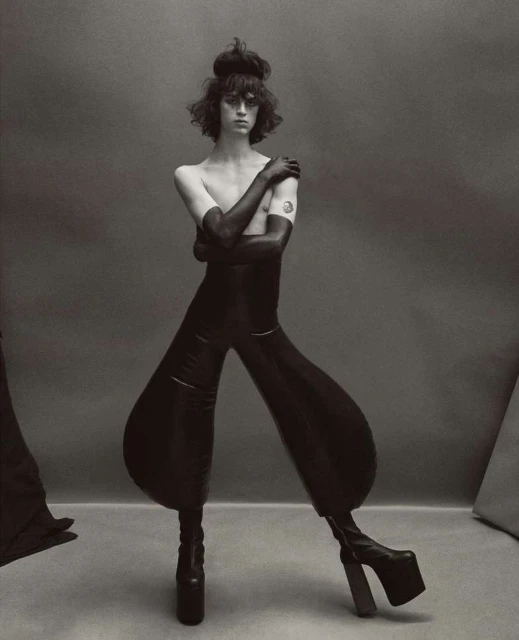 Black-and-white portrait of a person in a black bodysuit and platform boots posing in a studio against a neutral background.