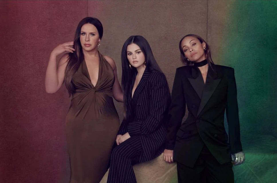 Three adult women pose indoors. One is seated in a pinstripe suit, and the other two are standing in an olive dress and a black suit.