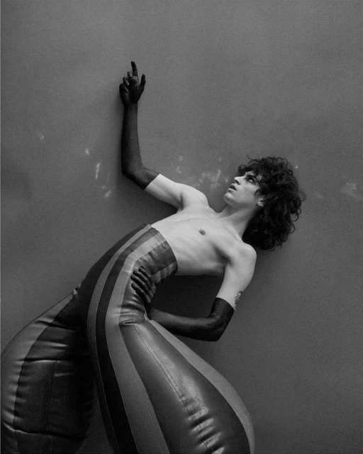 Avant-garde portrait of a bare-chested male fashion model in striped trousers and black gloves, posing against a solid grey background.