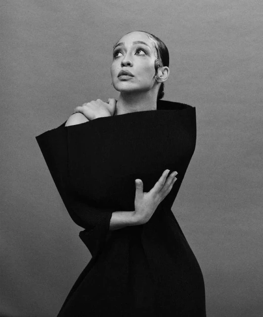 Elegant studio portrait of woman in a black sculptural dress, captured in monochrome with an upward gaze and hands gently framing her.